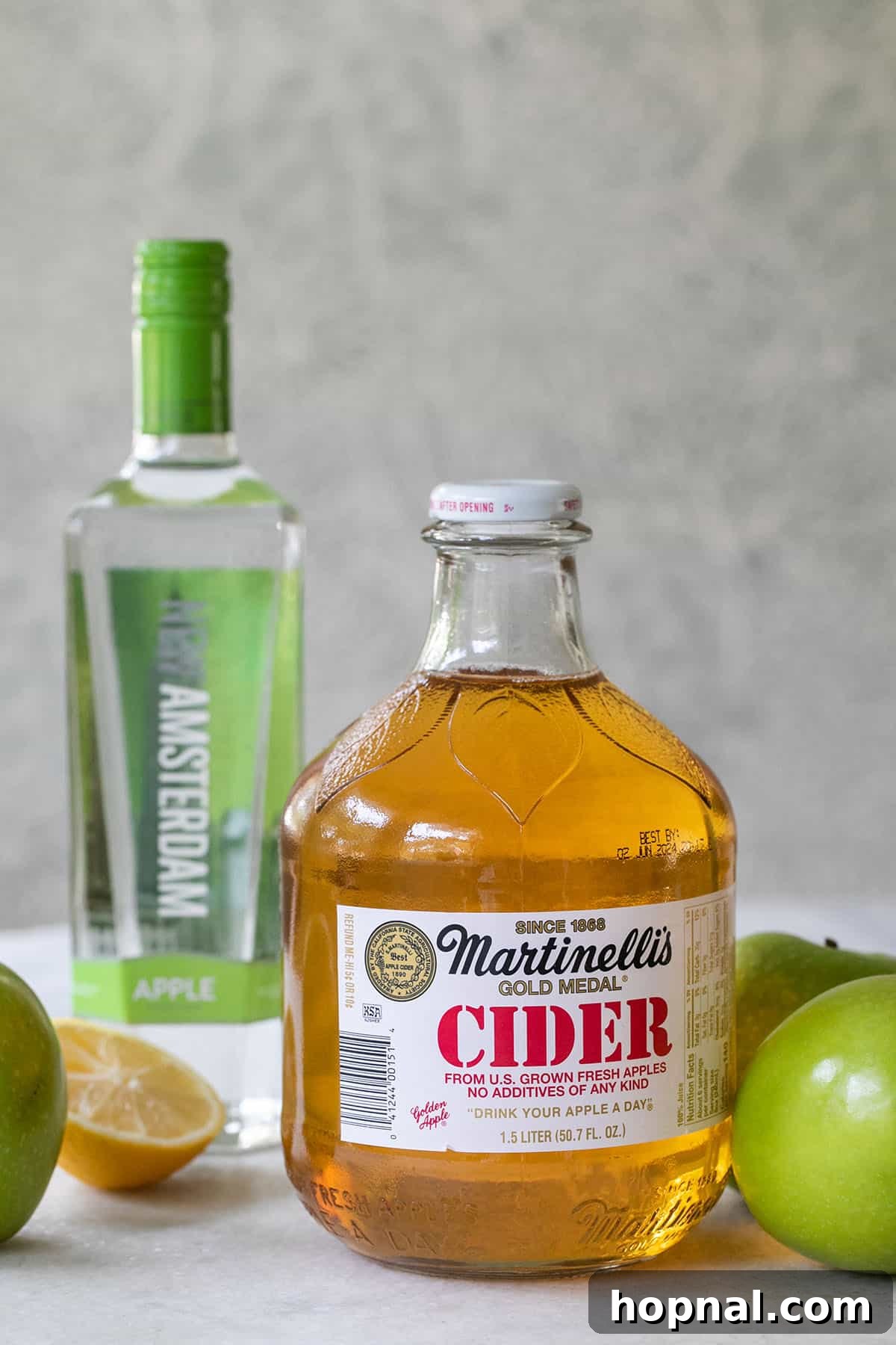 The three essential ingredients for an Apple Martini: a bottle of premium apple vodka, a glass of fresh apple cider, and a sliced lemon, all arranged artfully for a crisp cocktail.