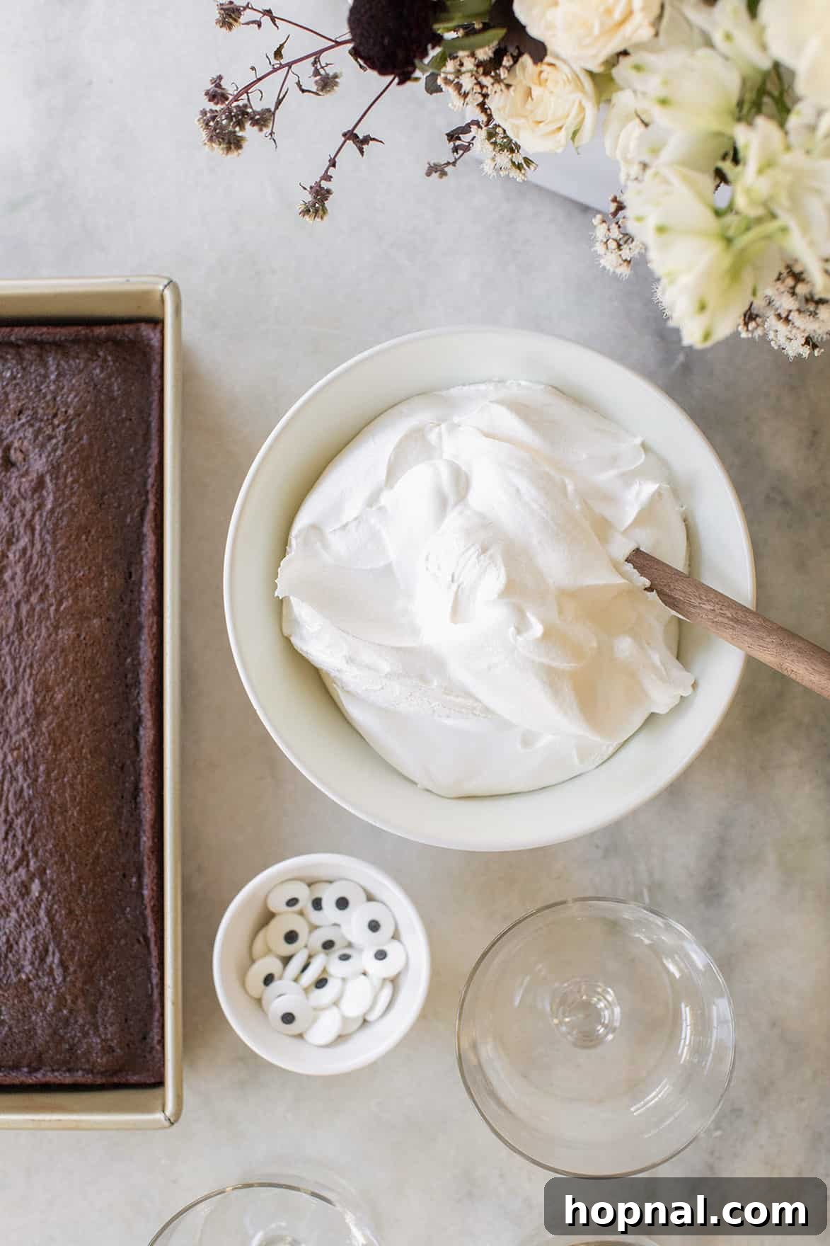 Ingredients for ghost chocolate trifles arranged on a wooden surface, including a bowl of fluffy whipped cream and a tray of freshly baked chocolate cake, ready for assembly