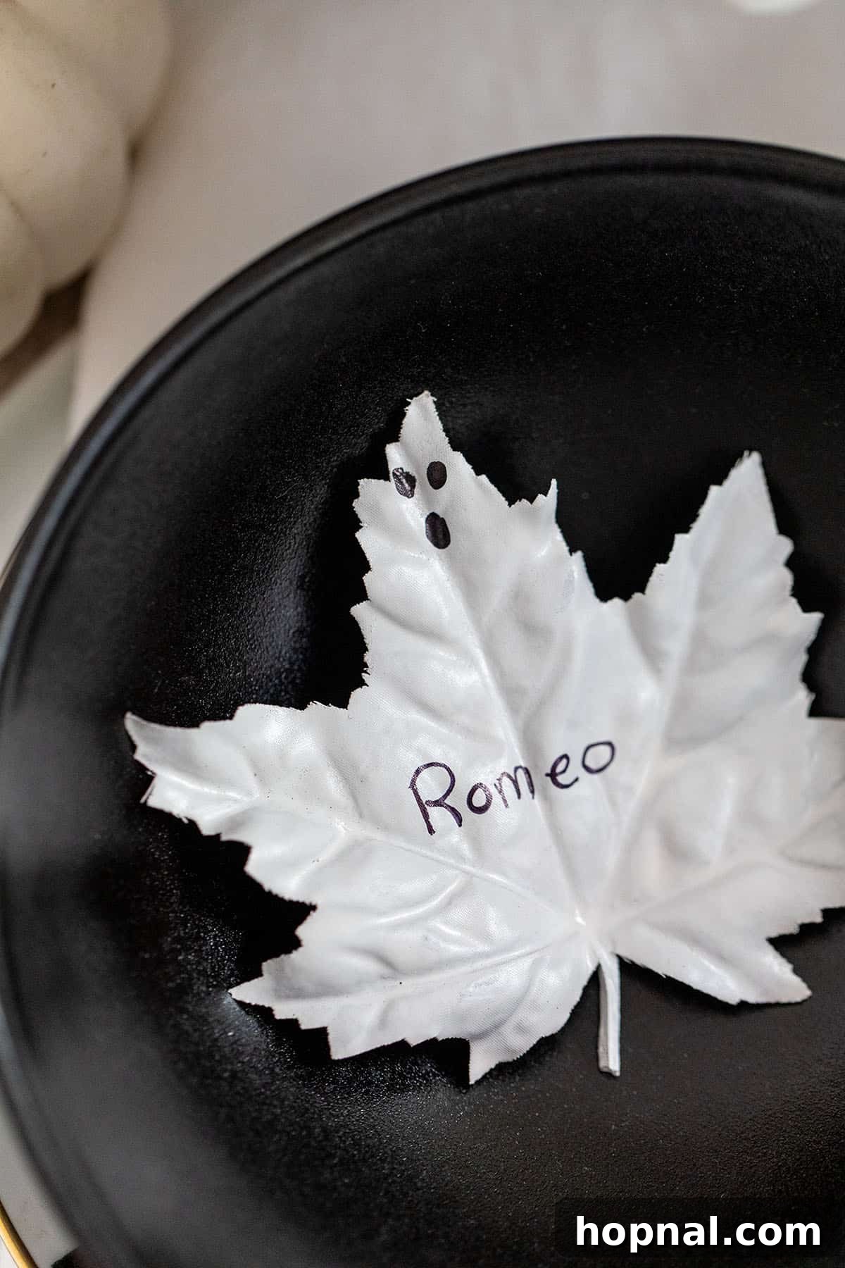 A Halloween seating card made from a white-painted maple leaf with a name and ghost face.