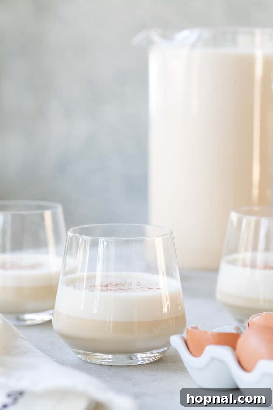 Rich and creamy homemade eggnog, spiked with bourbon, served in elegant glasses on a rustic wooden table alongside fresh eggs and whole spices.