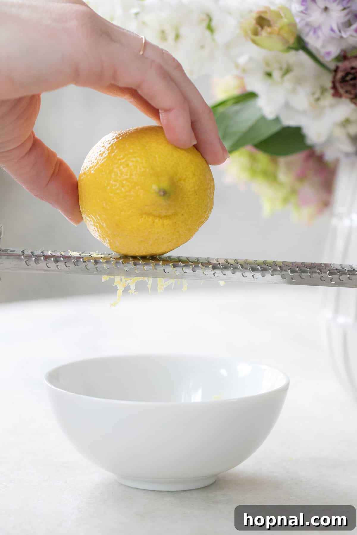 Close-up of a hand zesting a lemon with a microplane, showing the fine strands of yellow zest being produced over a bowl.