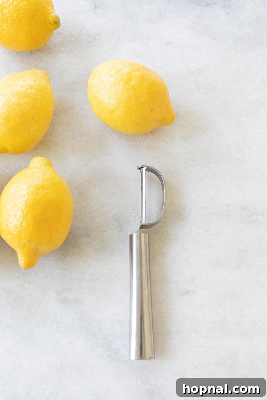 A vegetable peeler rests next to several fresh lemons, illustrating an alternative method for removing lemon peel for zest.