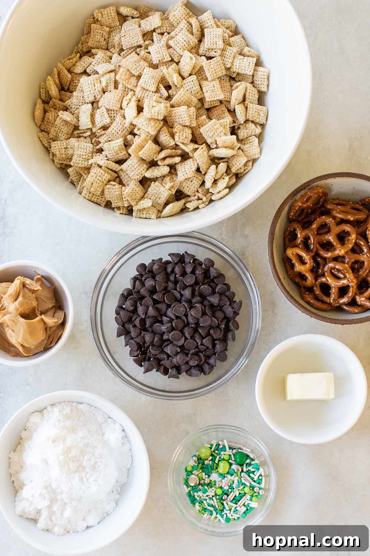 Magic Reindeer Dust 3 Various ingredients for Christmas Reindeer Food, including Rice Chex cereal, chocolate chips, mini pretzels, butter, and powdered sugar, neatly arranged in small bowls.