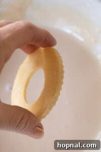 A hand dipping a baked wreath cookie into a bowl of vanilla glaze.