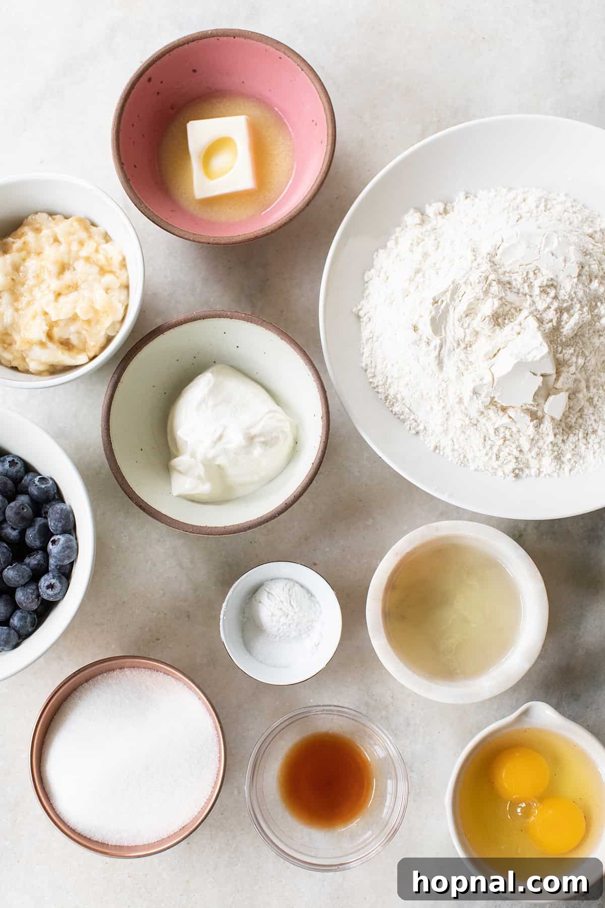 Golden Banana Blueberry Treats 4 Bowls of essential ingredients for blueberry banana muffins: eggs, flour, butter, sugar, vanilla, oil, sour cream, and fresh blueberries.