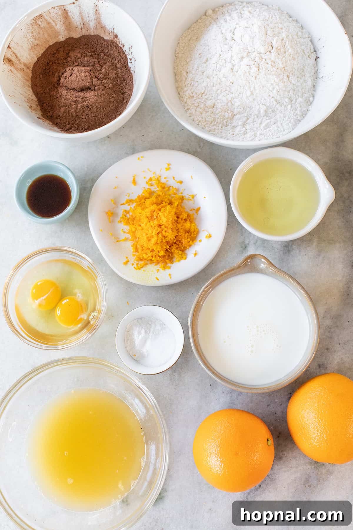 A vibrant flat lay of key chocolate orange cake ingredients including cocoa powder, fresh oranges, eggs, sugar, and flour.