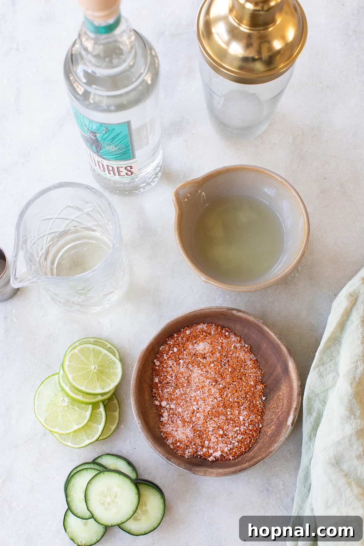 Fresh ingredients laid out: lime juice, crisp cucumbers, Tajín seasoning, and a bottle of tequila, alongside a cocktail shaker.