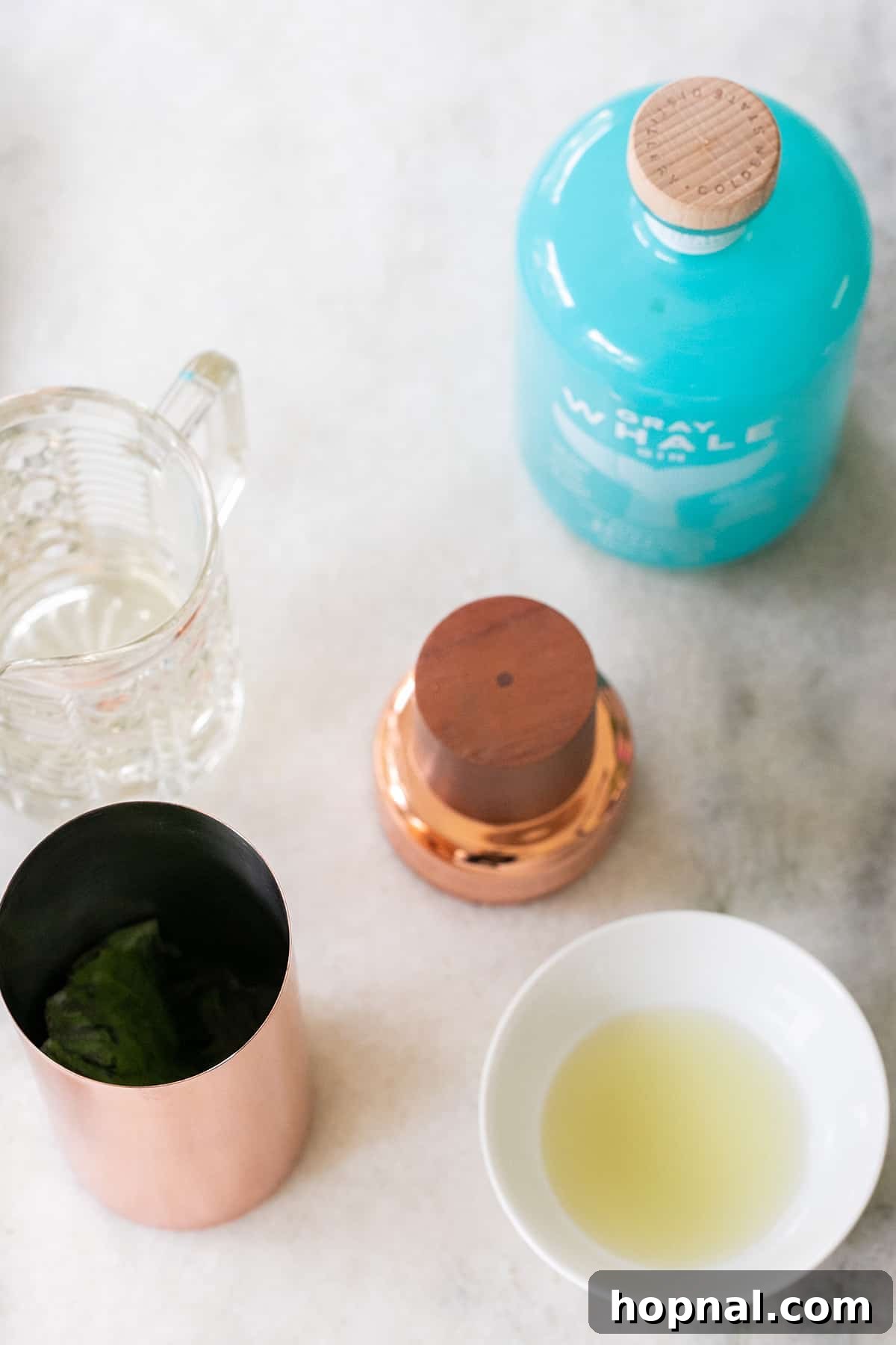 Essential ingredients for a Gin Basil Smash: gin bottle, fresh limes, cocktail shaker, and simple syrup on a rustic table.