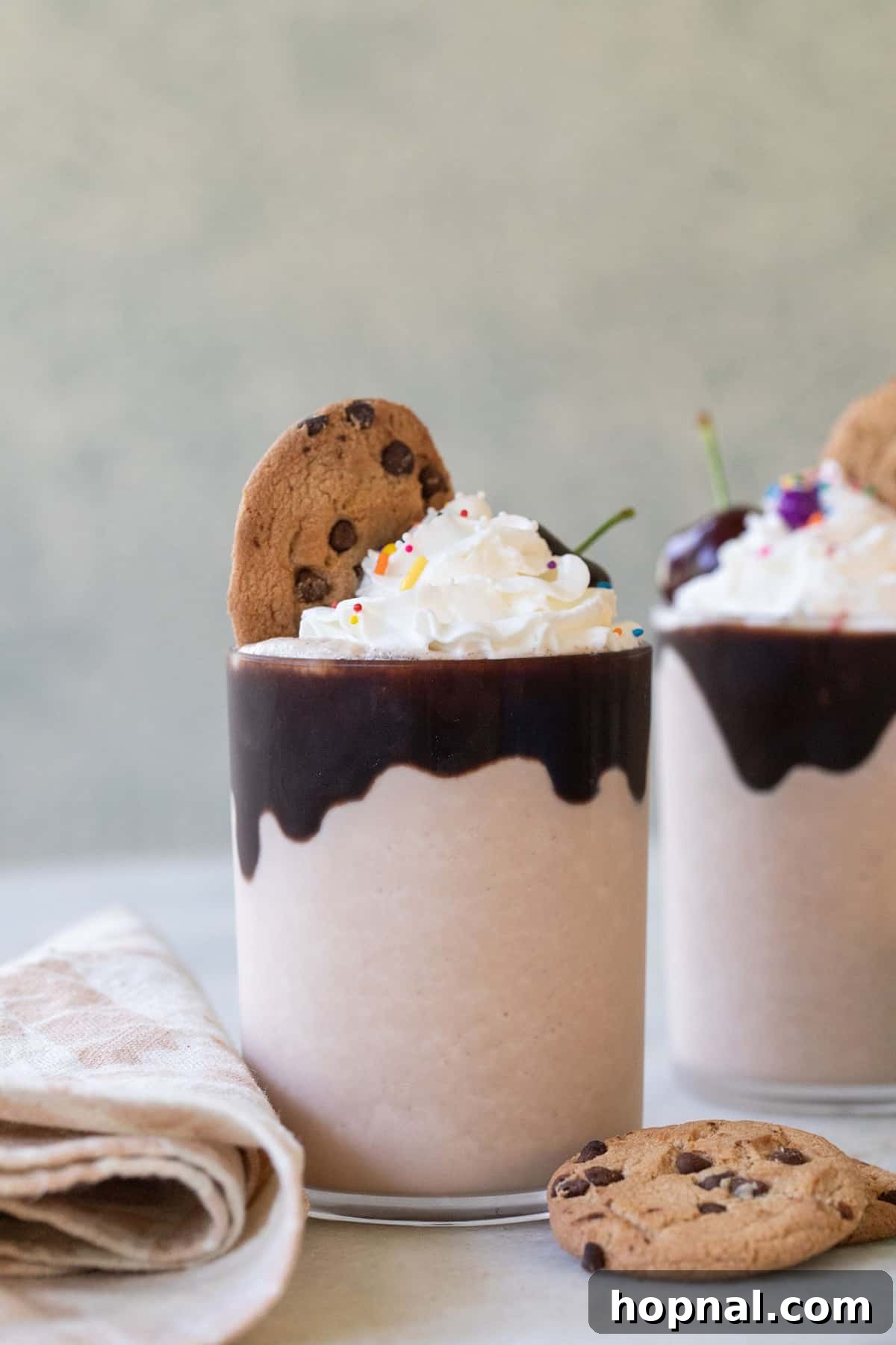 A tall glass filled with a creamy Chips Ahoy milkshake, drizzled with chocolate sauce, topped with a generous swirl of whipped cream, and garnished with crumbled cookies and a whole Chips Ahoy cookie. The background suggests a cozy setting, inviting you to enjoy this delicious chocolate chip cookie milkshake.
