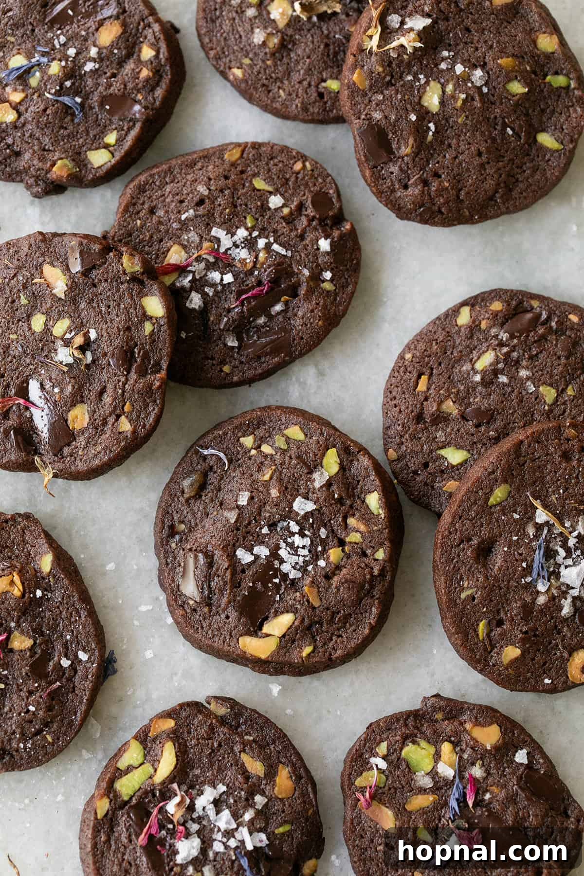 Exquisite chocolate sable cookies adorned with pistachios, flaky sea salt, and delicate edible flowers, arranged on a rustic wooden surface.