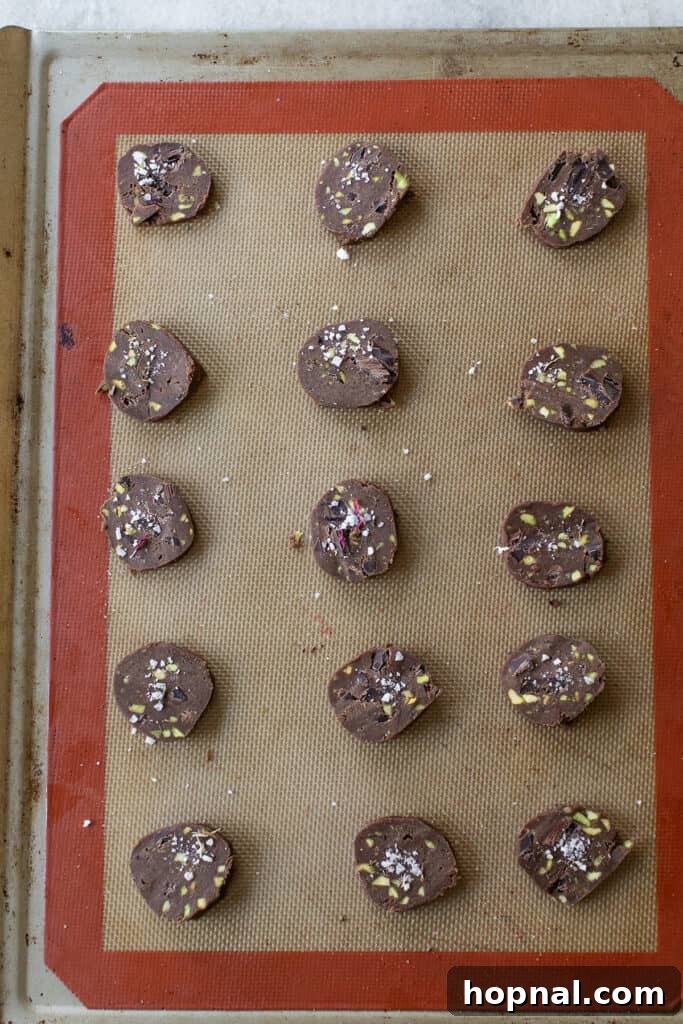 Unbaked chocolate pistachio sable cookies, perfectly sliced and arranged on a silpat-lined baking sheet, awaiting their transformation in the oven.
