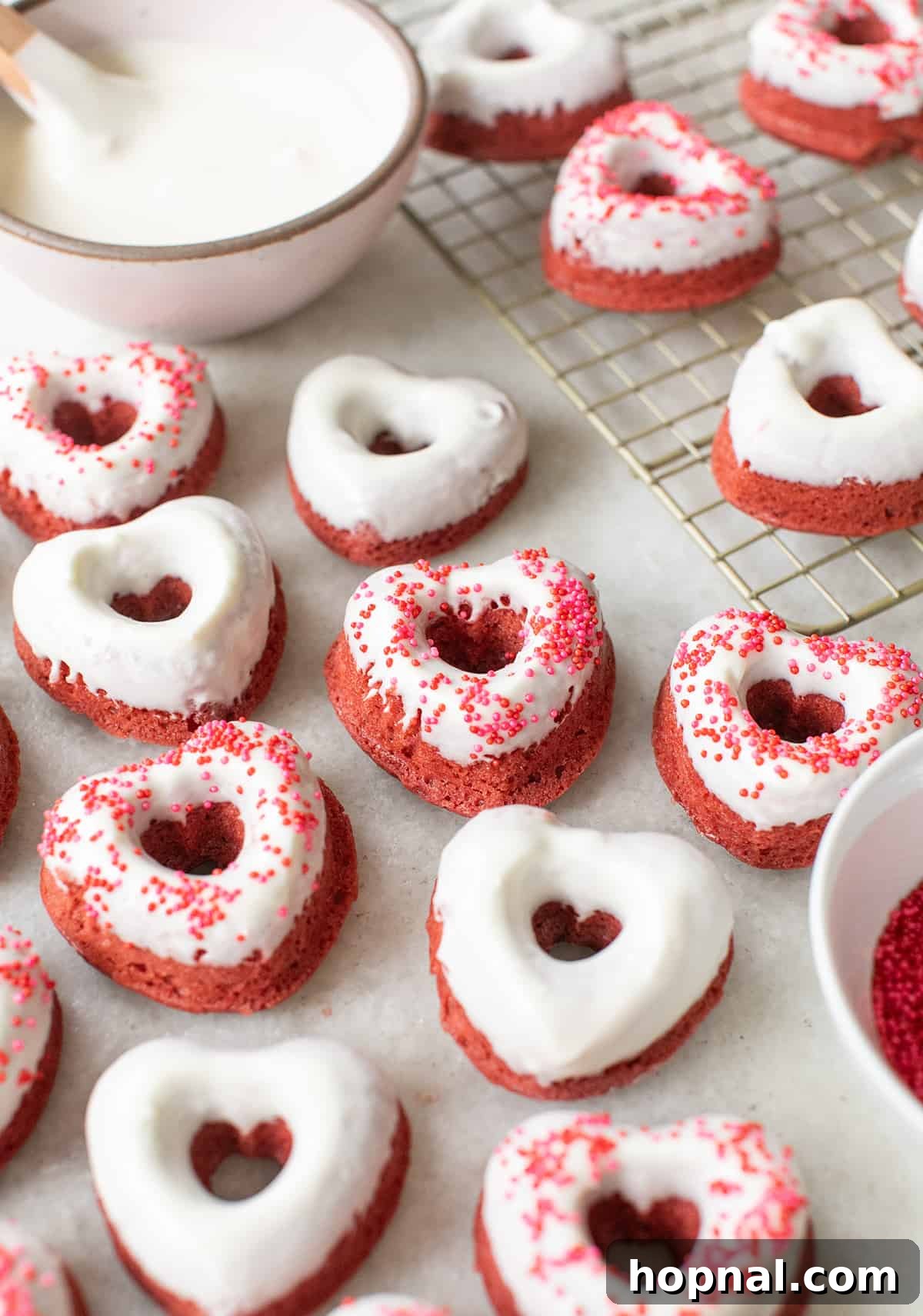 A collection of heart-shaped red velvet cake donuts, elegantly arranged on a serving plate, each glistening with cream cheese glaze and a sprinkle of decorative toppings.
