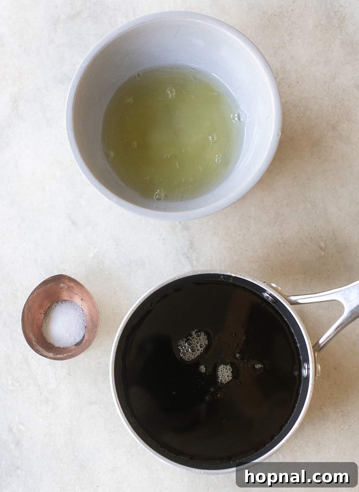 Three ingredients for maple frosting: fresh egg whites in a bowl, a bottle of pure maple syrup, and a small pinch of salt on a marble counter.