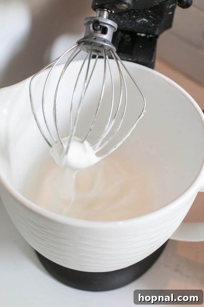 Egg whites being vigorously whipped in a stand mixer with the whisk attachment, forming soft peaks.