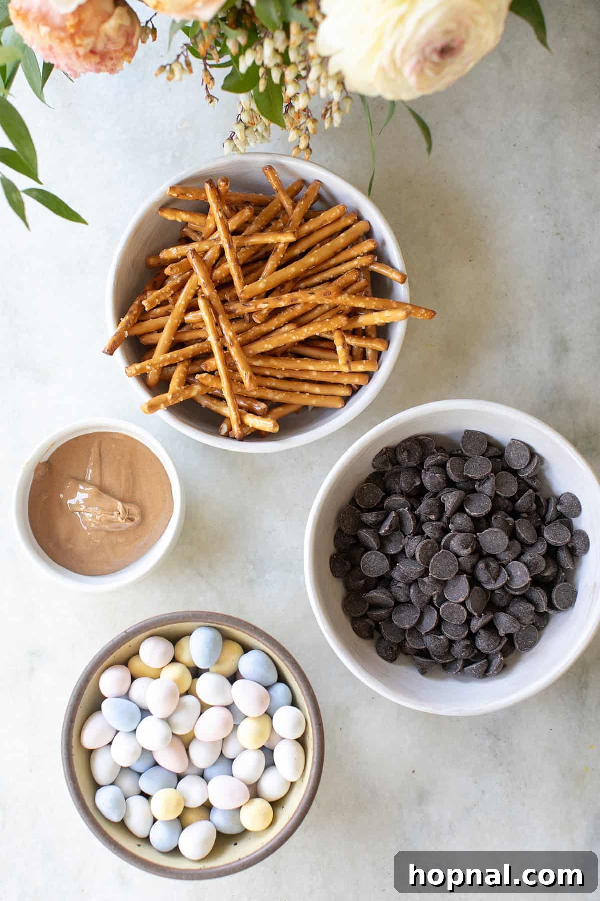 Sweet Spring Nests 4 The four essential ingredients for Bird's Nest Cookies laid out: small pretzel twists, semi-sweet chocolate chips, a jar of peanut butter, and colorful candy-coated chocolate eggs.