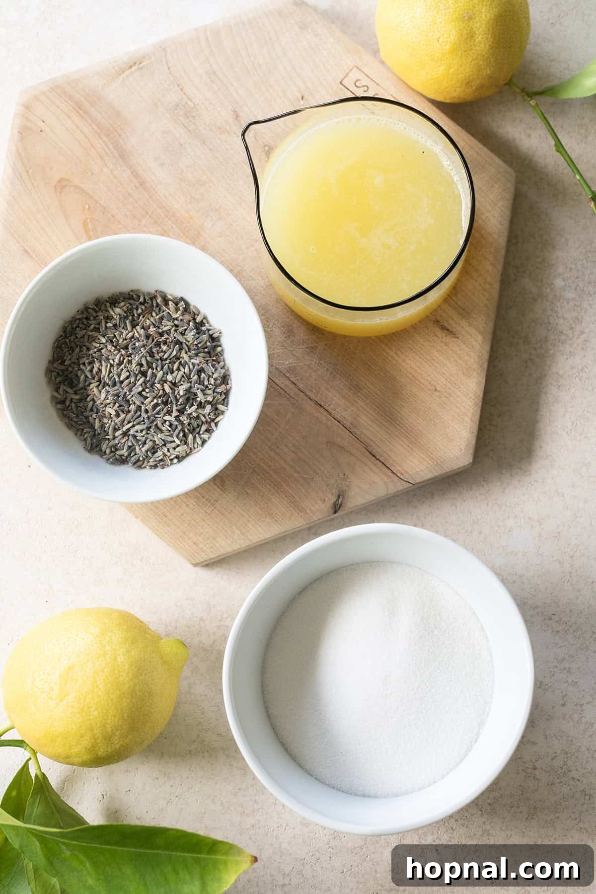 Fresh lemons, granulated sugar, dried culinary lavender, and a glass of water, laid out as ingredients for lavender lemonade.